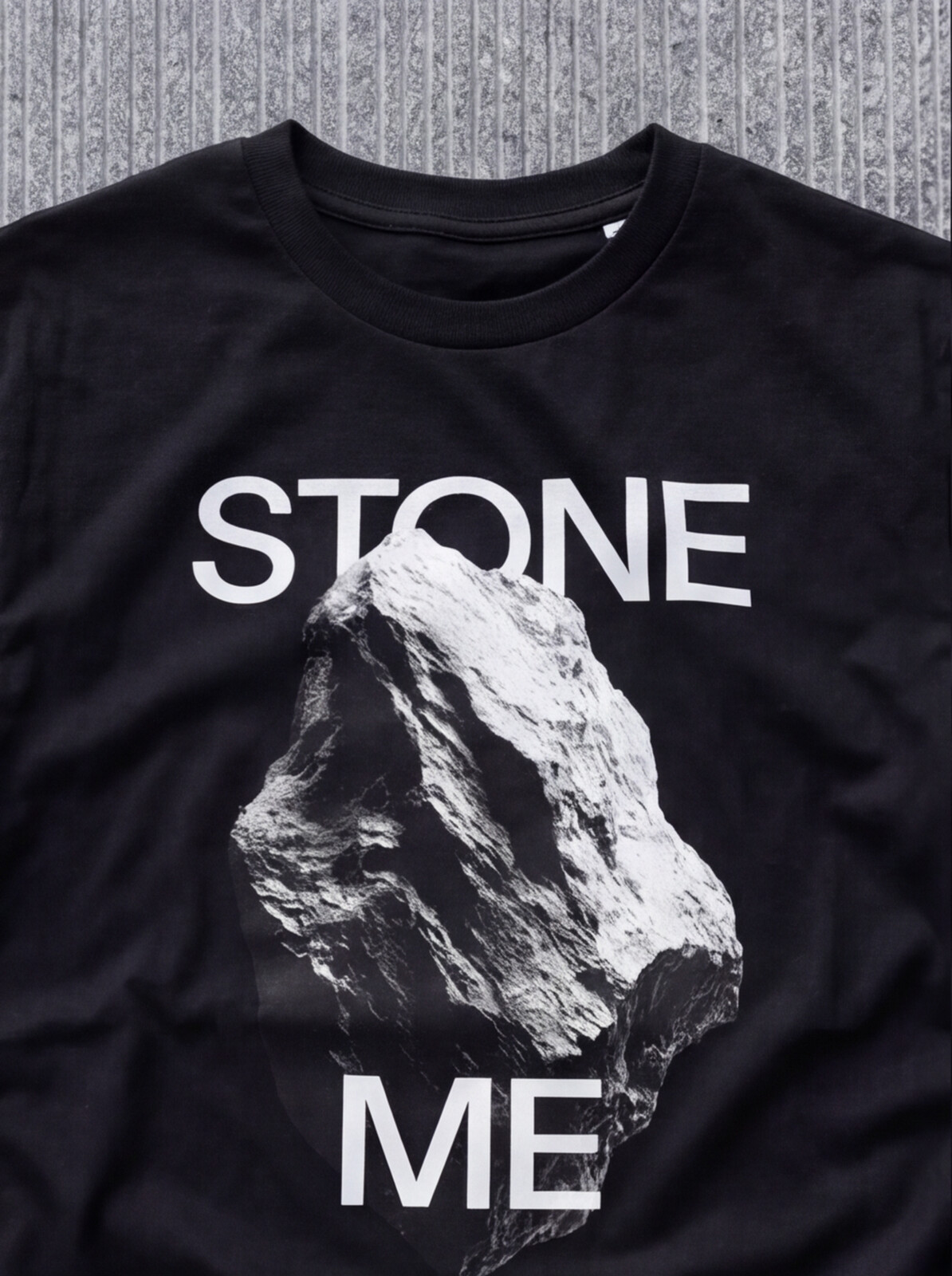 STONE ME tee from The Fifth State of Matter series — black t-shirt on concrete, front view